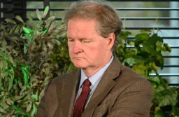 Prof. Grzegorz Kucharczyk z opuszczonym wzrokiem, w brązowej marynarce i bordowym krawacie, na tle zieleni i żaluzji