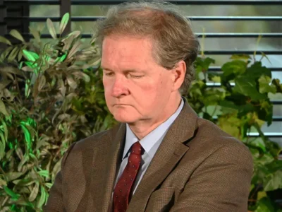 Prof. Grzegorz Kucharczyk z opuszczonym wzrokiem, w brązowej marynarce i bordowym krawacie, na tle zieleni i żaluzji