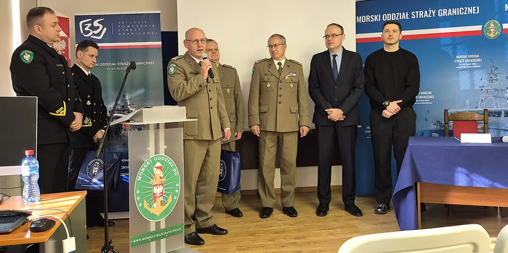 Mężczyzna w mundurze Straży Granicznej przemawia przy mównicy Morskiego Oddziału SG, obok stoją oficerowie i goście