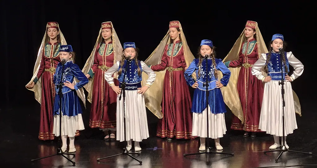 Dziewczęta w tatarskich strojach, w czerwonych sukniach ze złotymi welonami i niebieskich kamizelkach, na scenie przy mikrofonach