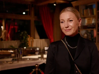 Uśmiechnięta kobieta w czarnym żakiecie i złotym łańcuszku we wnętrzu restauracji z czerwonymi zasłonami