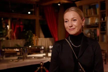 Uśmiechnięta kobieta w czarnym żakiecie i złotym łańcuszku we wnętrzu restauracji z czerwonymi zasłonami