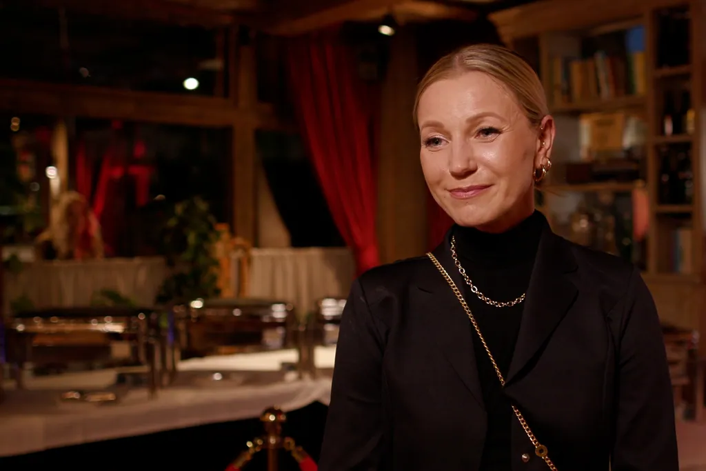 Uśmiechnięta kobieta w czarnym żakiecie i złotym łańcuszku we wnętrzu restauracji z czerwonymi zasłonami