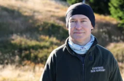 Mężczyzna w czapce i polarze z napisem Karkonoski Park Narodowy na tle górskiej łąki