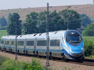 Biało-niebieski pociąg Pendolino PKP Intercity jedzie po torach wśród drzew i pól